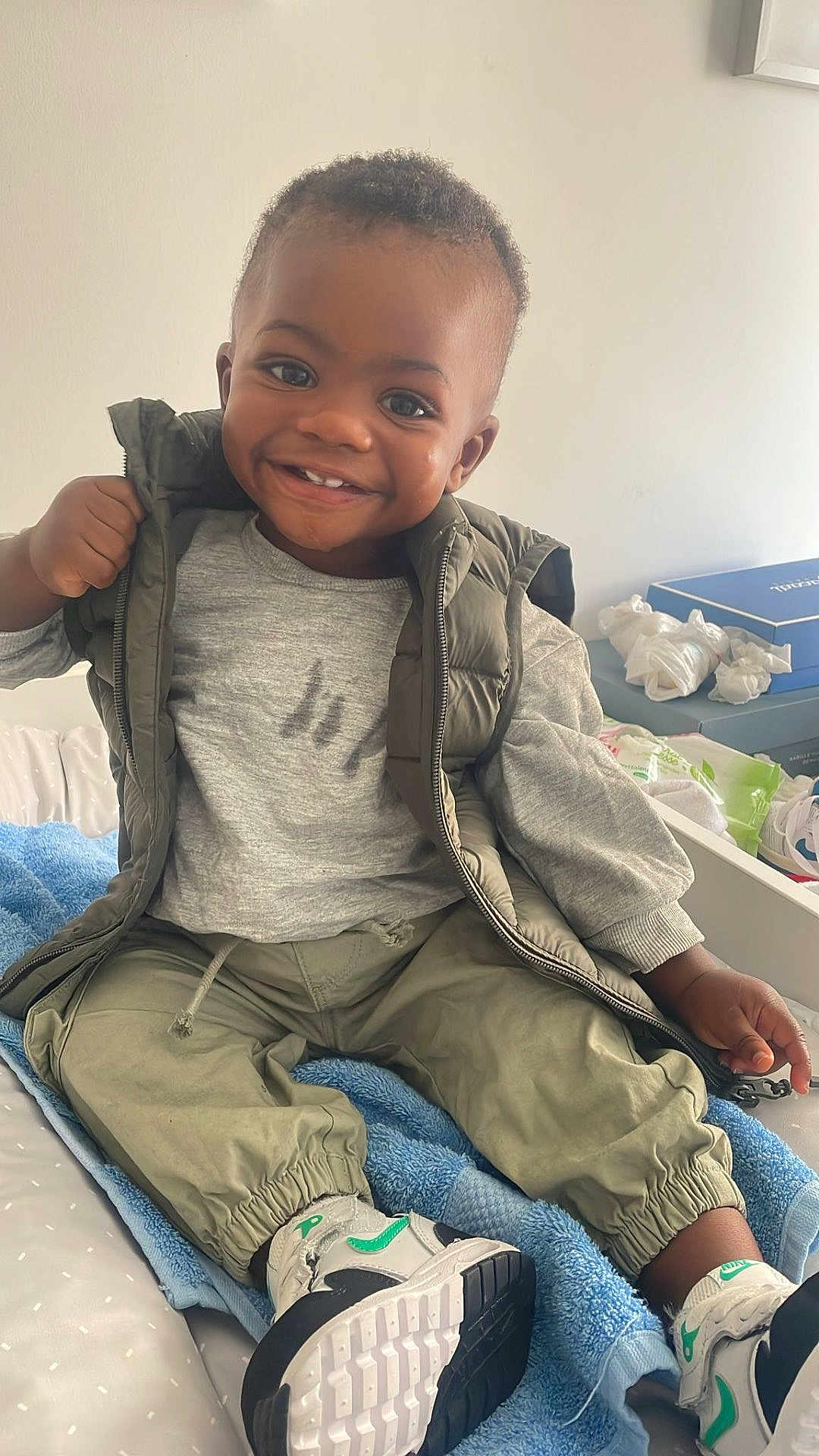 Ysaac a rejoint le concours — aidez-le/la à gagner de superbes lots ! bed, blanket, blue_blanket, casual, child, clothing, drawer, face, furniture, gray_shirt, green_vest, hair, happy, indoor, pants, person, smile, sneakers, toddler, white_blanket