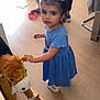 blue_dress, casual_clothing, child, curious, floor, furniture, hair_ties, indoor, looking_up, person, pigtails, playroom, rocking_horse, soft_toy, standing, toddler, toy, white_shoes, wooden_floor, young_girl