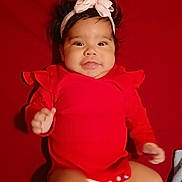 Jessica joined the competition — help win amazing prizes! baby, infant, child, smiling, red_onesie, headband, pink_bow, diaper, lying_down, red_background, bed, blanket, chubby_cheeks, dark_hair, portrait, cute, hands, eyes_open, indoor, soft_lighting