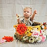 child, toddler, baby, portrait, basket, flowers, daisies, roses, orange_flowers, floral, garden, wicker, sitting, blonde_hair, smiling, outdoor_style, studio_portrait, fabric_backdrop, colorful, holding_flower