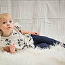 child, girl, toddler, portrait, indoors, sitting, lying_down, floral_dress, leggings, socks, blonde_hair, braids, blue_eyes, serious_expression, white_blanket, furry_rug, cushion, soft_lighting, studio_photo, childhood