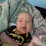 Thomas joined the competition — help win amazing prizes! baby, infant, batman_onesie, blanket, cozy, smiling, cute, child, soft, comfort, portrait, indoor, clothing, person, happy, young, resting, baby_hand, head, face