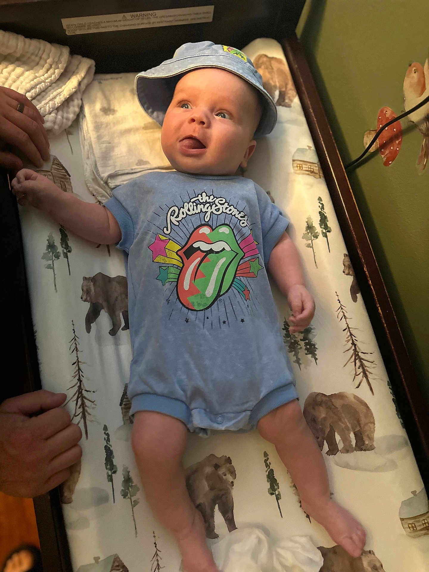 Thomas joined the competition — help win amazing prizes! baby, infant, onesie, hat, tongue_out, changing_pad, bear_pattern, hand, adult_hand, blue_clothing, baby_boy, indoor, cute, playful, expression, child, person, smiling, face, headwear
