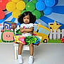 Amiri is registered to the contest to win money with this photo: child, girl, colorful_skirt, white_shoes, balloons, birthday_backdrop, cartoon, yellow_bus, bench, crossed_arms, expression, indoor, decorations, playful, cute, curly_hair, serious_face, party, celebration, toy