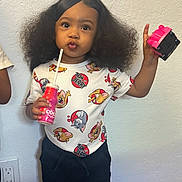 Amiri is registered to the contest to win money with this photo: toddler, child, indoor, standing, toy, drink, pink, shirt, cartoon, tom_and_jerry, black_pants, curly_hair, voluminous_hair, wall, floor, footwear, expression, person, young_child, casual_clothing