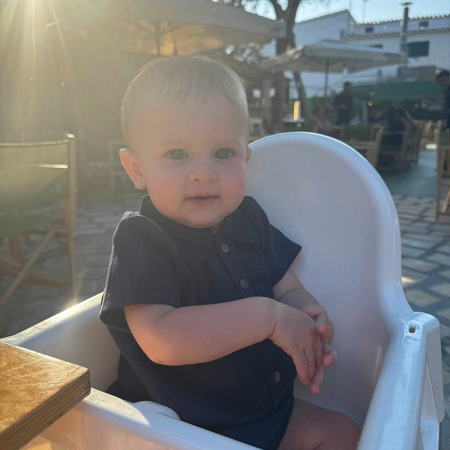 Tommy is registered to the contest to win money with this photo: baby, child, clothing, cute, daylight, face, happy, high_chair, infant, outdoor, person, restaurant, seat, skin, summer, sunflare, sunlight, table, trees, umbrella