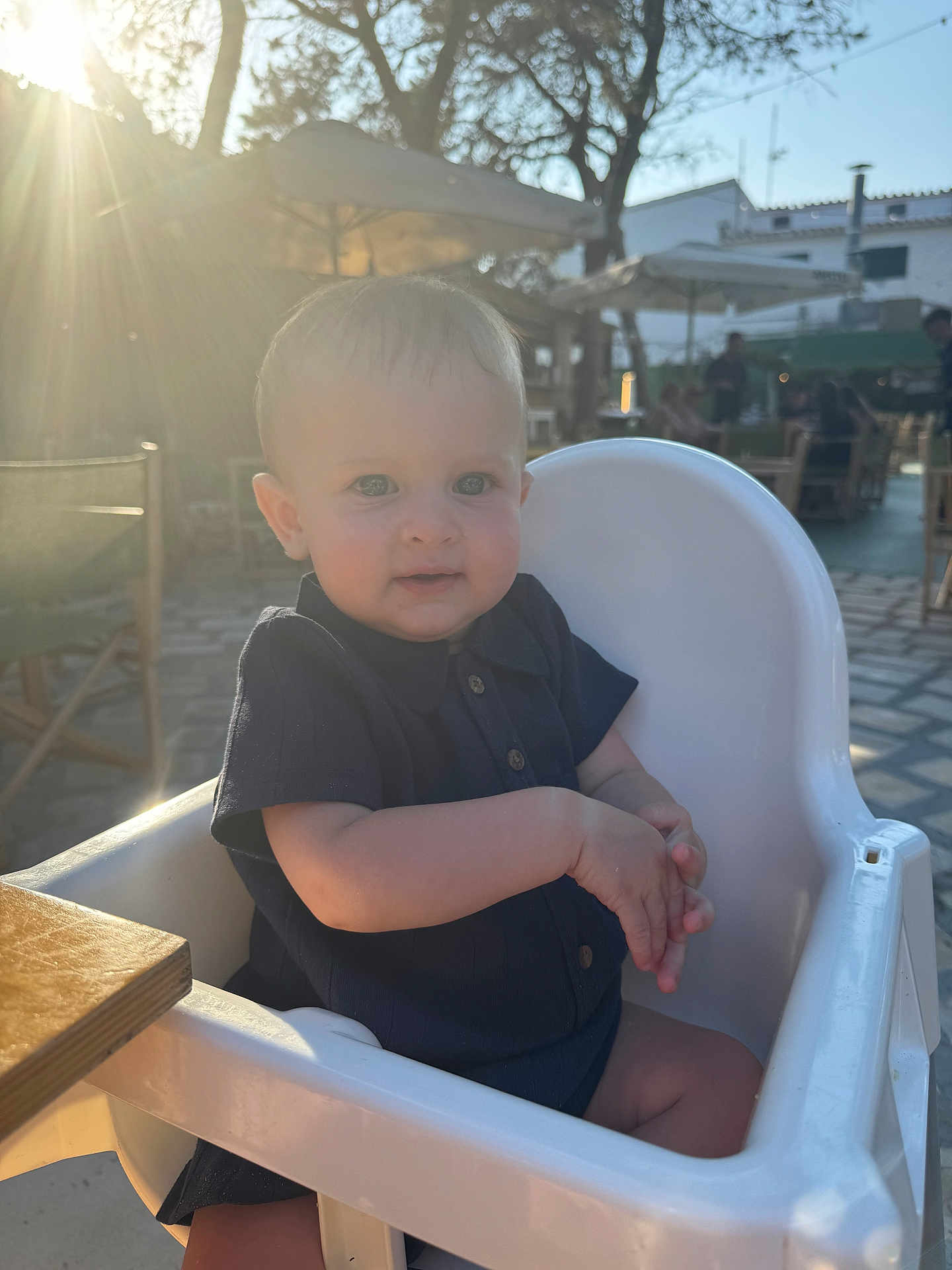 Tommy is registered to the contest to win money with this photo: baby, child, high_chair, outdoor, sunlight, sunflare, trees, umbrella, table, clothing, person, skin, face, seat, summer, daylight, restaurant, cute, infant, happy