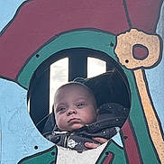 Junior is registered to the contest to win money with this photo: baby, child, face, portrait, circle_cutout, painted_board, outdoor, colorful, expressionless, person, jacket, hand, background, art, playground, window, wood, sky, casual, young