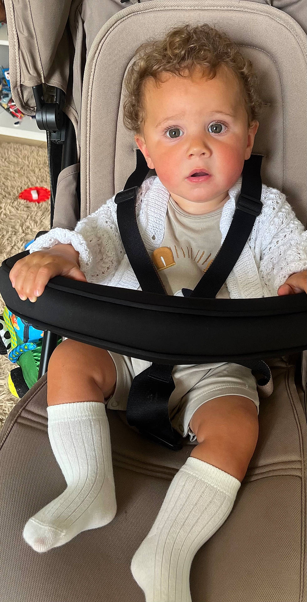 Freddie is registered to the contest to win money with this photo: baby, beige_clothing, candid, carpet, child, curious_expression, curly_hair, face, footwear, hand, indoor, knitted_cardigan, legs, person, portrait, seatbelt, stroller, toddler, toy, white_socks