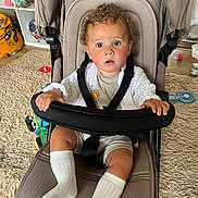 Freddie joined the competition — help win amazing prizes! toddler, child, stroller, curly_hair, indoor, carpet, toy, white_socks, clothing, seatbelt, expression, baby, hand, face, curious, furniture, floor, room, person, sitting