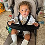 toddler, child, stroller, curly_hair, indoor, carpet, toy, white_socks, clothing, seatbelt, expression, baby, hand, face, curious, furniture, floor, room, person, sitting
