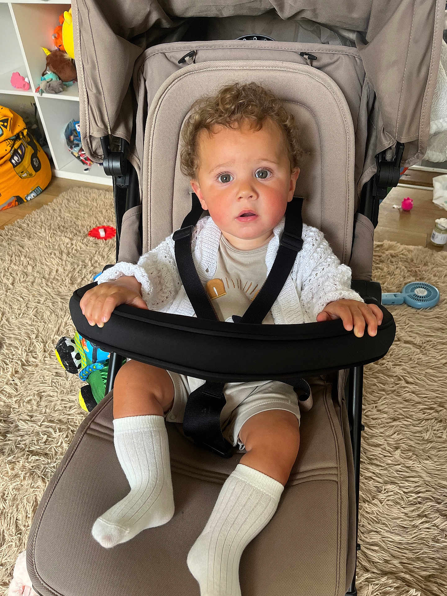 Freddie joined the competition — help win amazing prizes! toddler, child, stroller, curly_hair, indoor, carpet, toy, white_socks, clothing, seatbelt, expression, baby, hand, face, curious, furniture, floor, room, person, sitting