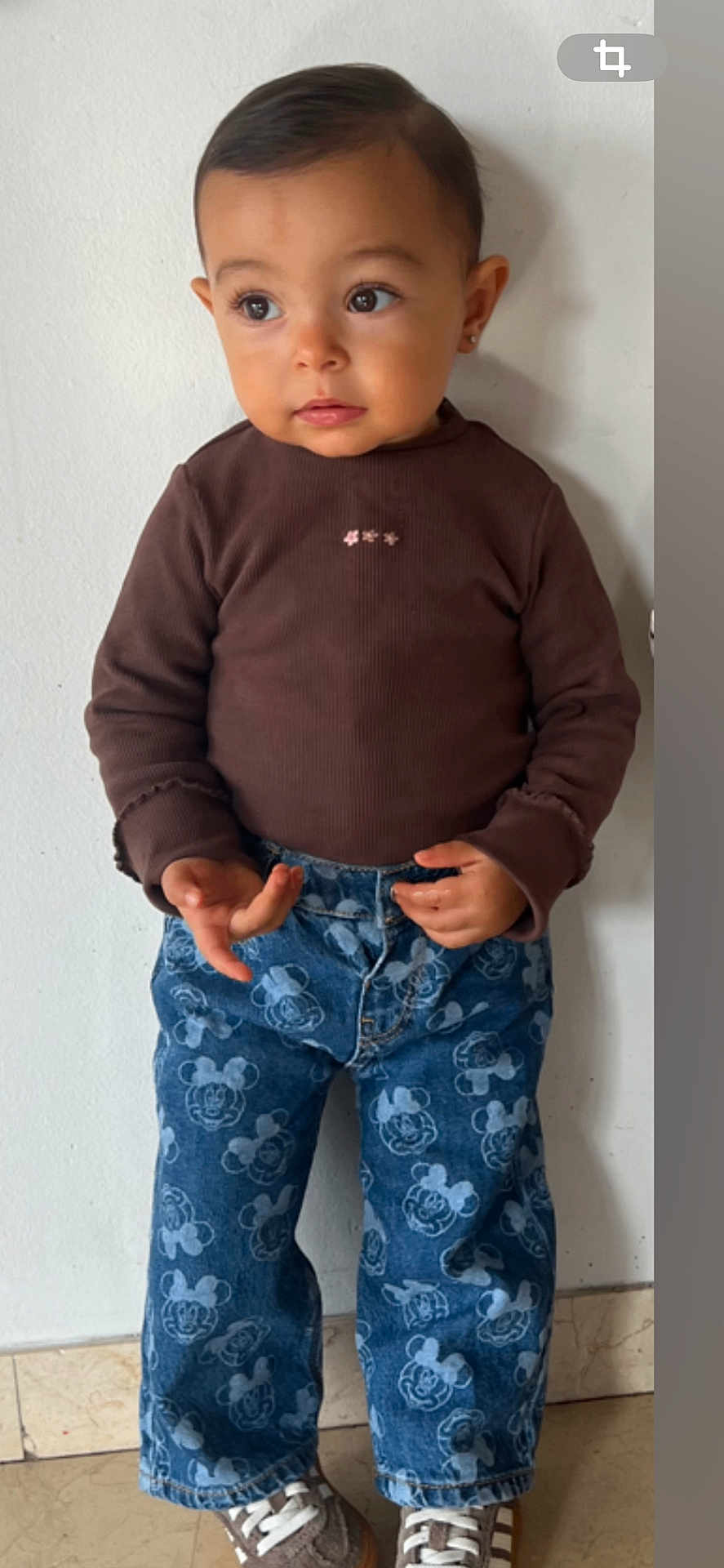 Yéva participe au concours pour gagner de l'argent avec cette photo : toddler, child, standing, brown_shirt, blue_jeans, mickey_mouse, curly_hair, earrings, sneakers, indoor, wall, floor, casual_clothing, cute, small_hands, portrait, young_child, fashion, person, curious