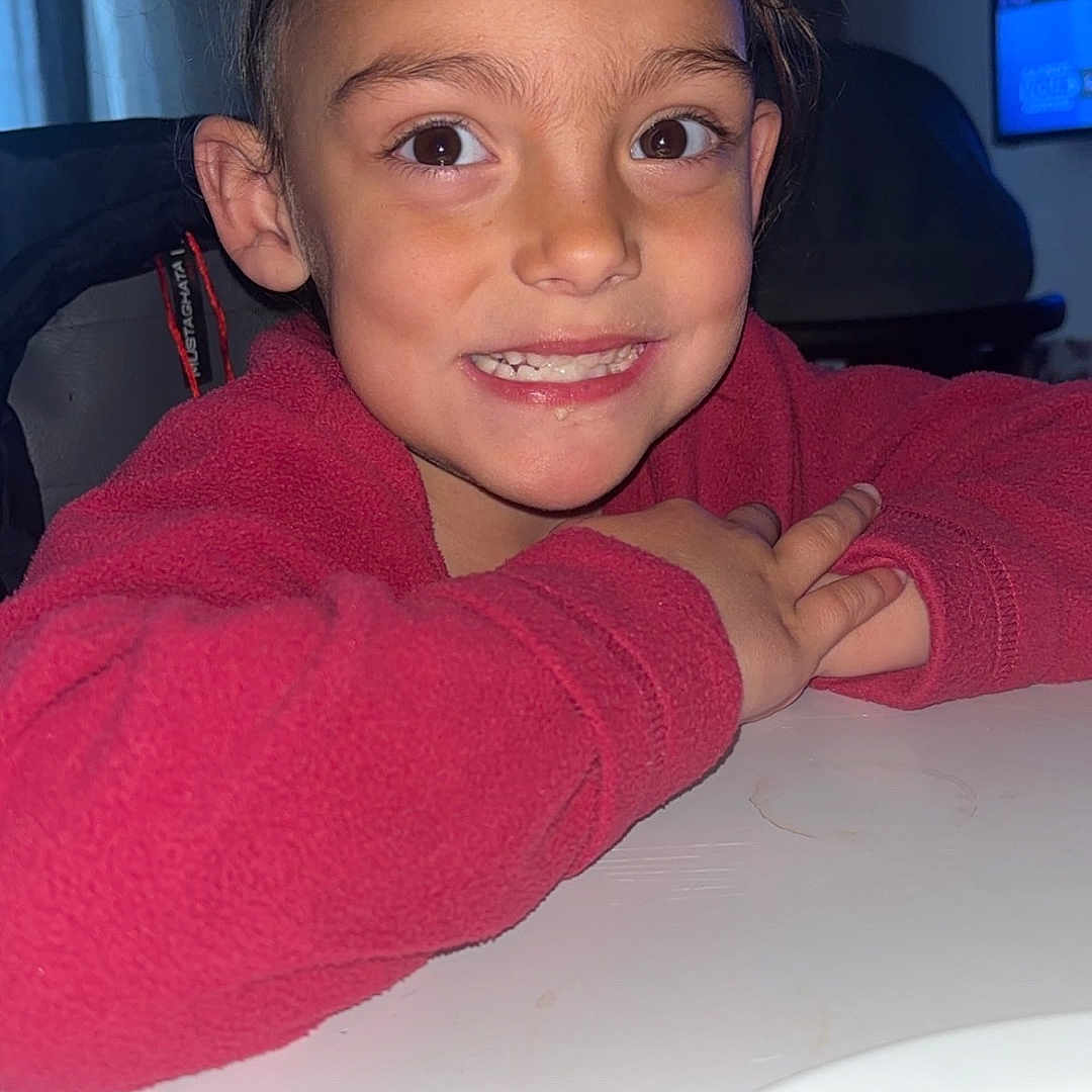 Sayana participe au concours pour gagner de l'argent avec cette photo : child, smiling, face, red_sweater, table, indoor, portrait, young_child, happy, person, cozy, curtains, television, brown_hair, casual, candid, home, expression, light, room