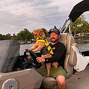 Penny is registered to the contest to win money with this photo: man, dog, boat, life_jacket, water, lake, nature, outdoor, trees, sky, cloudy, smiling, hat, casual_clothing, steering_wheel, seat, recreation, summer, adventure, pet
