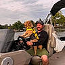 man, dog, boat, life_jacket, water, lake, nature, outdoor, trees, sky, cloudy, smiling, hat, casual_clothing, steering_wheel, seat, recreation, summer, adventure, pet