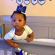 Taytum is registered to the contest to win money with this photo: baby, child, crib, crown, banner, tassels, celebration, half_birthday, infant, smiling, face, hand, white_clothing, bedroom, furniture, decor, indoor, cute, curious, portrait