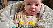 Zoe is registered to the contest to win money with this photo: baby, baby_seat, blanket, child, colorful, cozy, cute, expression, face, home, indoor, infant, kitchen, person, playful, refrigerator, soft_texture, tongue_out, toy, wooden_cabinets