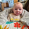 Zoe is registered to the contest to win money with this photo: baby, infant, blanket, face, tongue_out, baby_seat, toy, colorful, indoor, kitchen, wooden_cabinets, refrigerator, soft_texture, expression, child, cute, playful, person, home, cozy