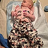 baby, infant, clothing, pink_top, cow_print_pants, lying_down, changing_pad, blanket, foot, hand, face, person, indoor, curious, soft_texture, blue_cup, skin, casual, newborn, portrait