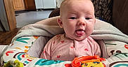 Zoe is registered to the contest to win money with this photo: baby, infant, baby_seat, toy, blanket, tongue_out, indoor, kitchen, furniture, colorful, person, cute, child, playful, wood_floor, cabinet, refrigerator, table, window, lighting
