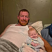 Zoe joined the competition — help win amazing prizes! man, baby, smile, blanket, couch, tank_top, relaxing, indoor, wall, red_hair, beard, tattoo, pillow, comfort, family, portrait, resting, happy, person, casual