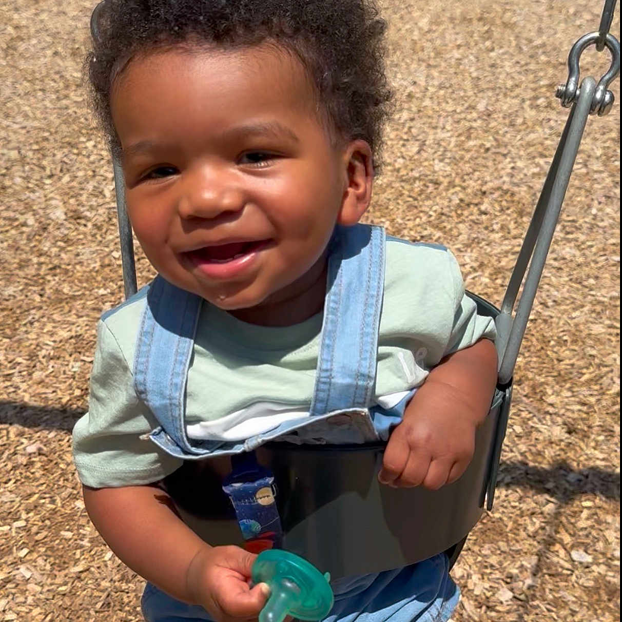 Marcal joined the competition — help win amazing prizes! baby, child, clothing, curly_hair, denim_overalls, fun, green_pacifier, happy, outdoor, pacifier, person, play, playground, shorts, smiling, summer, sunlight, swing, toddler, wood_chips