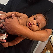 Isaiah is registered to the contest to win money with this photo: baby, infant, diaper, arm, hand, person, indoor, curious, looking_at_camera, black_shirt, skin, close_up, holding, home, kitchen, furniture, adult, face, human, portrait