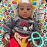 baby, bouncer, child, colorful, comfort, curly_hair, cute, expression, face, hand, indoor, infant, mickey_mouse, patterned_background, red_pants, seated, sweatshirt, teething_toy, toy, young