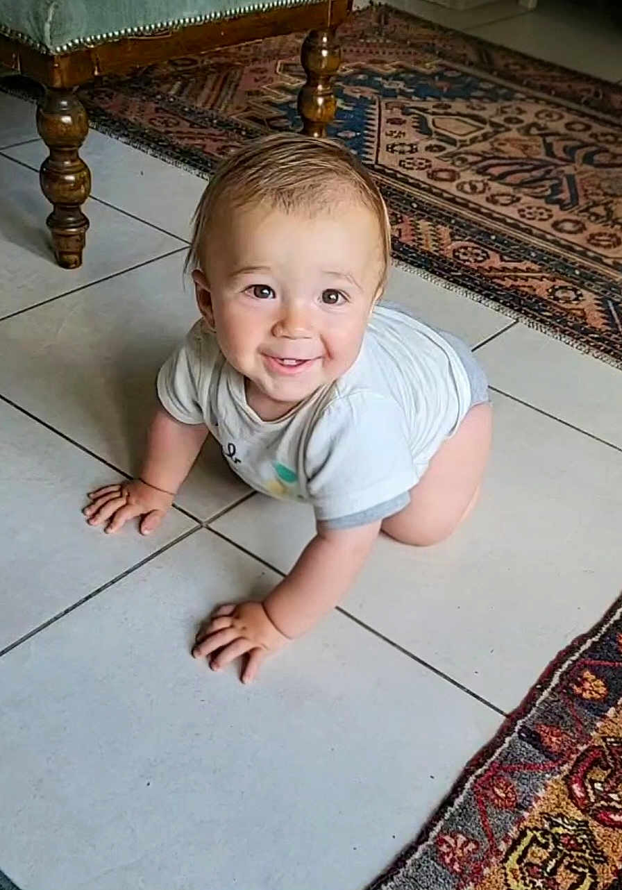 Hugo participe au concours pour gagner de l'argent avec cette photo : baby, crawling, floor, tiled_floor, rug, patterned_rug, wooden_furniture, smiling, happy, indoor, child, cute, person, short_hair, white_shirt, gray_shorts, hands, eyes, face, home