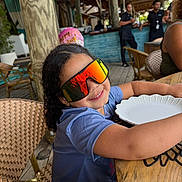 Maia Nunez is registered to the contest to win money with this photo: child, curly_hair, sunglasses, smile, plate, wooden_table, restaurant, balloon, casual_clothing, indoor, chair, person, happy, reflection, glasses, table, celebration, people, background, food_service