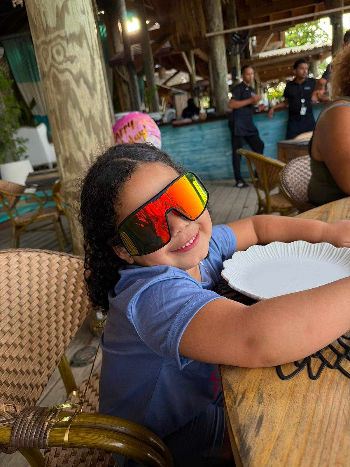 Maia Nunez is registered to the contest to win money with this photo: child, curly_hair, sunglasses, smile, plate, wooden_table, restaurant, balloon, casual_clothing, indoor, chair, person, happy, reflection, glasses, table, celebration, people, background, food_service