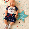 4th_of_july, baby, blue_shorts, celebration, clothing, confetti, cute, festive, fluffy_blanket, happy, holiday, independence_day, infant, laying_down, portrait, red_white_and_blue, sign, smiling, star_pattern, white_shoes
