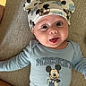 apparel, baby, blue_clothing, child, cute, expression, face, happy, hat, headwear, indoor, infant, lying_down, mammal, mickey_mouse, onesie, person, portrait, smiling, soft_lighting