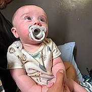 Apollo is registered to the contest to win money with this photo: baby, pacifier, adult_hand, beard, face, infant, holding, indoor, clothing, onesie, skin, person, expression, portrait, cute, child, head, human, parent, family