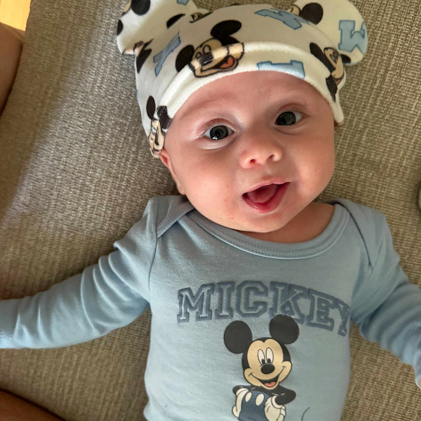 Apollo joined the competition — help win amazing prizes! baby, child, mickey_mouse, hat, blue_onesie, smiling, tongue_out, cute, infant, face, headwear, lying_down, cushion, fabric, happy, person, portrait, clothing, cartoon_character, indoor