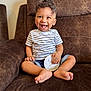toddler, child, smiling, sitting, striped_shirt, shorts, barefoot, couch, indoor, happy, curly_hair, milestone_card, brown_furniture, portrait, cute, young_child, casual, comfortable, baby, joyful