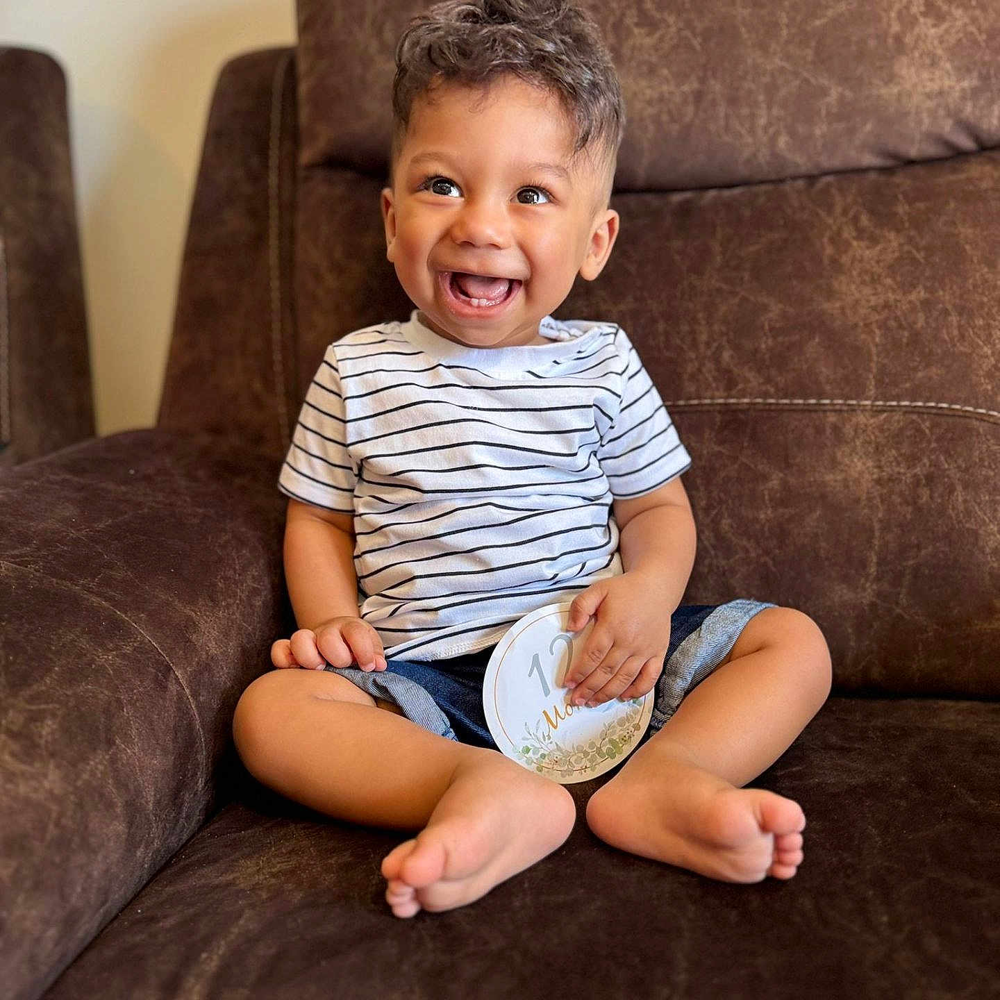 Zidan is registered to the contest to win money with this photo: baby, barefoot, brown_furniture, casual, child, comfortable, couch, curly_hair, cute, happy, indoor, joyful, milestone_card, portrait, shorts, sitting, smiling, striped_shirt, toddler, young_child