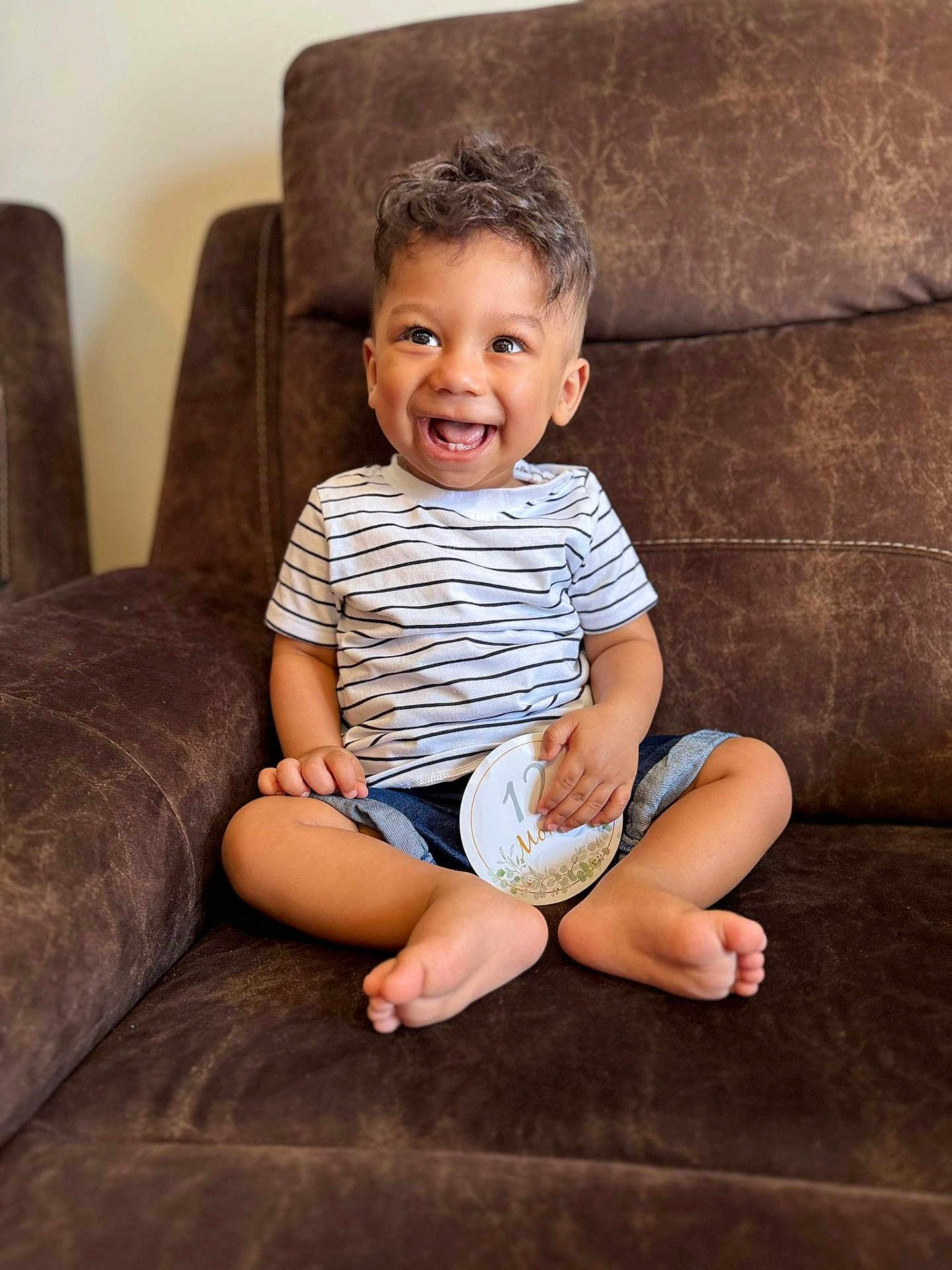 Zidan is registered to the contest to win money with this photo: toddler, child, smiling, sitting, striped_shirt, shorts, barefoot, couch, indoor, happy, curly_hair, milestone_card, brown_furniture, portrait, cute, young_child, casual, comfortable, baby, joyful