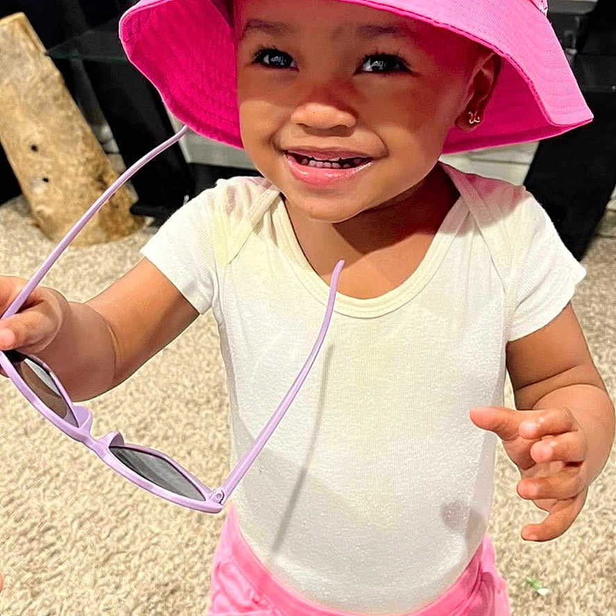 Jimmya is registered to the contest to win money with this photo: toddler, child, pink_bucket_hat, unicorn, flowers, sunglasses, white_tshirt, pink_shorts, smiling, standing, carpet, indoor, glass_table, plants, happy, cute, earrings, person, home, fashion