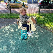 Agathe a rejoint le concours — aidez-le/la à gagner de superbes lots ! car, casual_clothing, child, curly_hair, daylight, fun, glasses, grass, green_fence, horse_toy, outdoor, pavement, play, playground, recycling_bins, shadow, smile, sneakers, spring_ride, tree