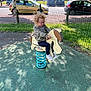 car, casual_clothing, child, curly_hair, daylight, fun, glasses, grass, green_fence, horse_toy, outdoor, pavement, play, playground, recycling_bins, shadow, smile, sneakers, spring_ride, tree