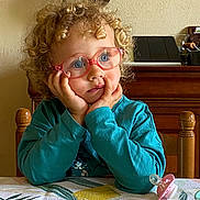 Agathe a rejoint le concours — aidez-le/la à gagner de superbes lots ! child, toddler, curly_hair, glasses, blue_eyes, table, tablecloth, pacifier, thoughtful, indoor, wooden_chair, wooden_furniture, portrait, sitting, cute, face, hands, clothing, long_sleeve, home