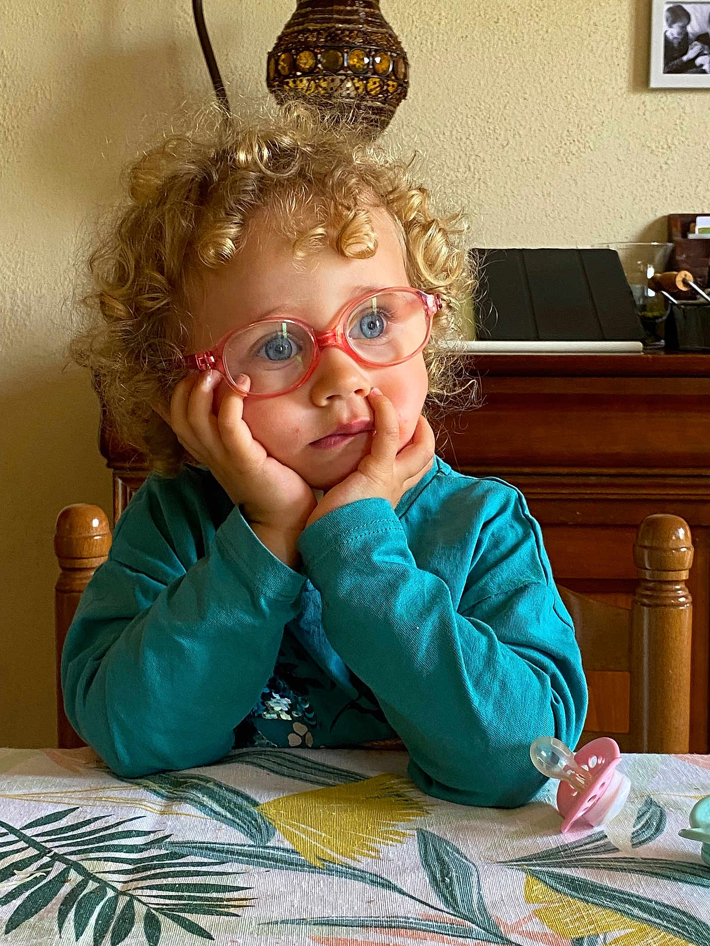 Agathe a rejoint le concours — aidez-le/la à gagner de superbes lots ! child, toddler, curly_hair, glasses, blue_eyes, table, tablecloth, pacifier, thoughtful, indoor, wooden_chair, wooden_furniture, portrait, sitting, cute, face, hands, clothing, long_sleeve, home