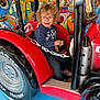 Agathe participe au concours pour gagner de l'argent avec cette photo : child, toy, tractor, amusement_ride, curly_hair, glasses, red, blue_floor, smiling, playful, indoor, person, seat, chain, colorful, background, vehicle, fun, young, happy