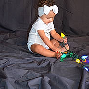Braylee joined the competition — help win amazing prizes! toddler, child, baby, headband, bow, lights, colorful_lights, sitting, curious, indoors, cloth_backdrop, wrinkled_cloth, playing, barefoot, white_onesie, portrait, young_child, toy, festive, cute