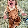 child, toddler, portrait, smiling, laughing, open_mouth, sitting, bed, bedding, plaid, overalls, corduroy, brown_clothing, blonde_hair, blue_eyes, indoors, water_bottle, playful, happy, face
