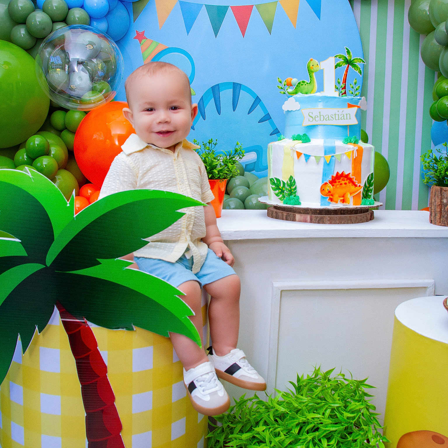 Sebastián is registered to the contest to win money with this photo: toddler, child, smile, birthday_cake, balloons, party, dinosaur, decorations, plant, table, shoes, shorts, shirt, smiling, indoor, celebration, colorful, props, happy, face