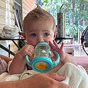 Charlie is registered to the contest to win money with this photo: baby, child, sippy_cup, blue, mohawk, towel, hand, porch, outdoor, sunlight, furniture, person, face, expression, holding, drink, table, background, skin, cute