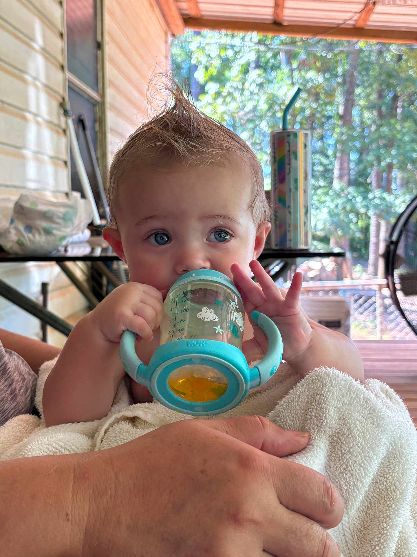 Charlie is registered to the contest to win money with this photo: baby, child, sippy_cup, blue, mohawk, towel, hand, porch, outdoor, sunlight, furniture, person, face, expression, holding, drink, table, background, skin, cute